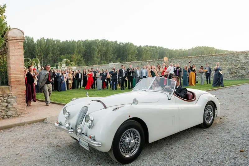 pareja de recién casados saliendo de la ceremonia de la boda en un antiguo y lujoso coche jaguar, los invitados aplauden al fondo mientras se van